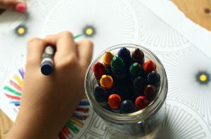 Kinderhand mit Wachsmalstift malt auf einem Stück Papier, dass auf einem Tisch liegt, daneben mehrere Wachsmalstifte in einem Glas