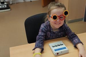 Mädchen mit Blümchen-Testbrille sitzt am Tisch vor einem kleinen Kästchen mit 4 Tasten