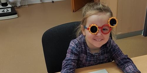 Mädchen mit Blümchen-Testbrille sitzt am Tisch vor einem kleinen Kästchen mit 4 Tasten